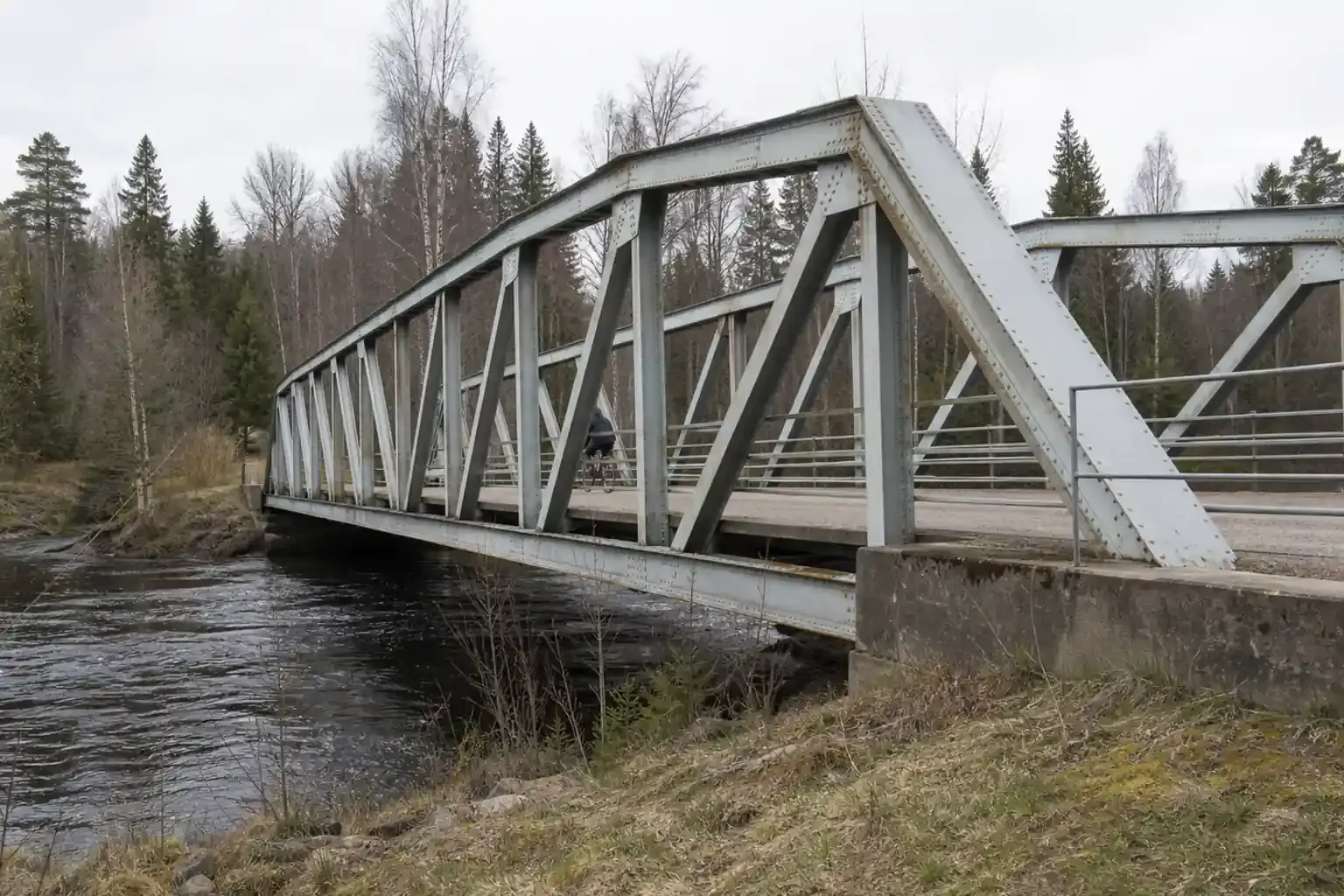 Fackverksbro av nitat stål över ett vattendrag i skogsmiljö